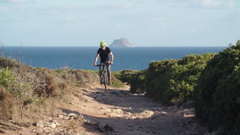 man rides mountain bike up dirt narrow rocky path in countryside by the coast, sardinia, italy, static