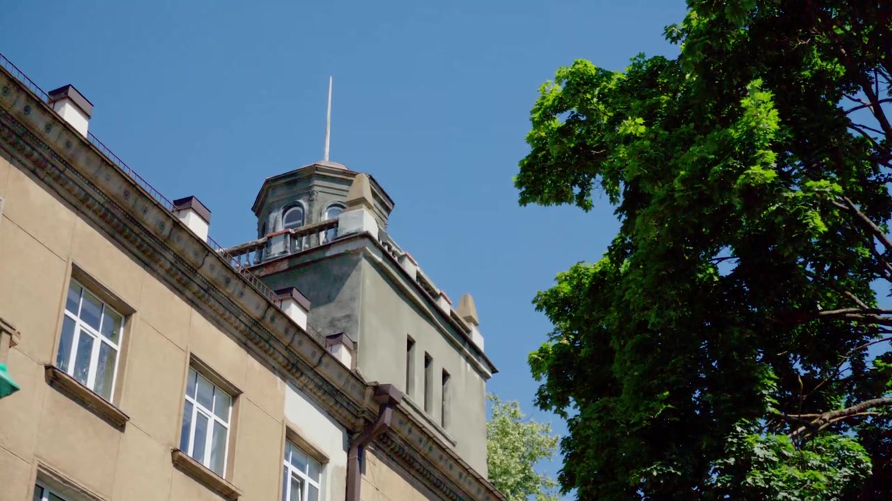 Inquadratura dal basso che mostra un angolo dettagliato della facciata con una torre a guglia decorativa su un edificio storico, incorniciato da un albero verde e frondoso, nella città di Daugavpils, Lettonia, in una giornata di sole.