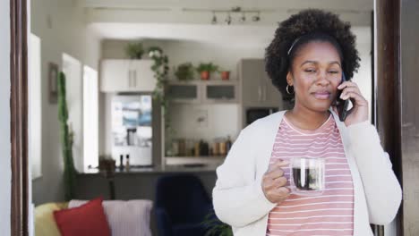 Portrait-of-happy-african-american-woman-drinking-tea-and-talking-on-smartphone-at-home