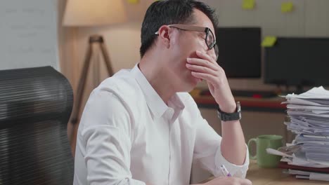 close up of tired asian man yawning and taking off his glasses before sleeping while working hard with documents at the office