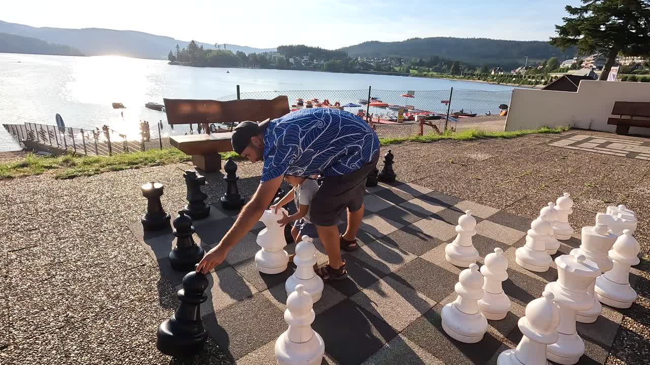 Pai e filho jogando xadrez gigante perto do lago