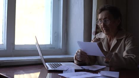 una anciana muy preocupada leyendo facturas en papel, pagando en línea en casa, administrando las finanzas del banco, calculando impuestos, tarifas, altas tasas de alquiler, planeando préstamos, deudas, pago de pensiones, sentada en la mesa de la cocina.