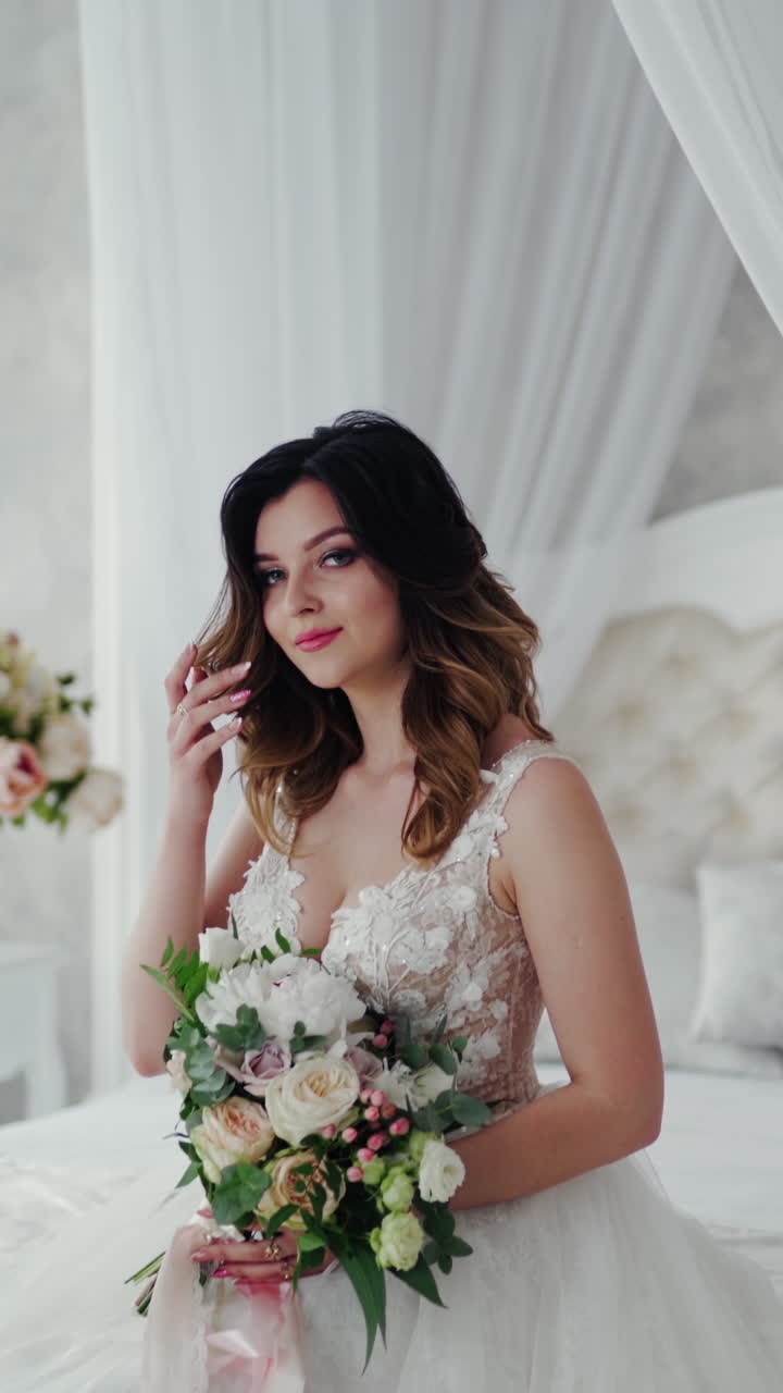 Beautiful young model in a white lace dress sits on a white bed. Gorgeous bride in wedding dress with flowers is sitting in the light bedroom. Vertical video