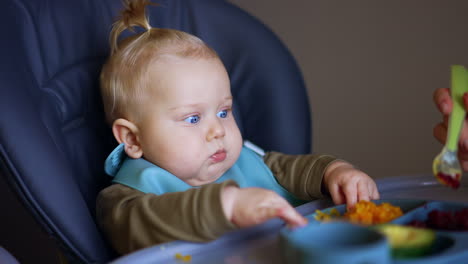 serious baby boy with a bib sits in high chair. mom gives boiled vegetables to her child.