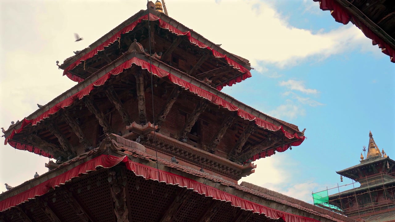 Et statisk mellembillede fra lav vinkel, der fokuserer på de indviklede tagkonstruktioner i flere niveauer af træ og røde udsmykninger på et gammelt hinduistisk pagodetempel mod en overskyet himmel i Nepal