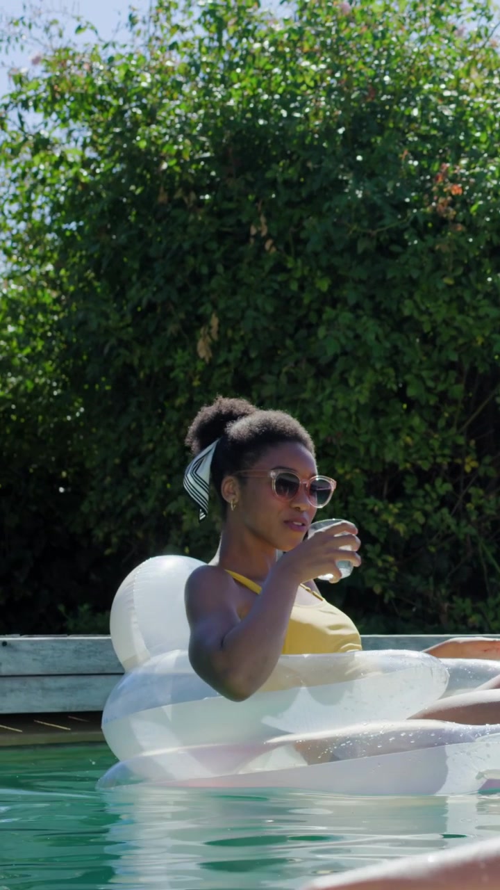 Vertical video: Panning camera to woman in yellow swim top and shades relaxing on float sipping cup