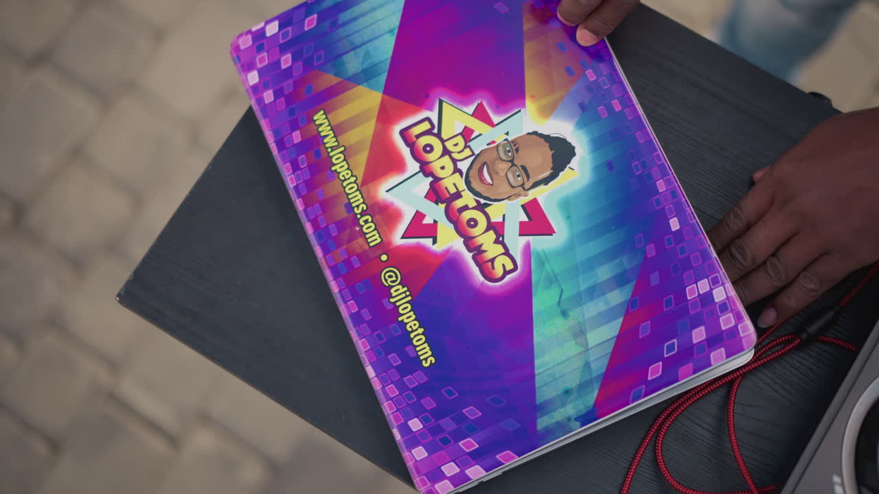 Cover colorata del laptop con tastiera arcobaleno, primo piano delle mani di un musicista di colore che sistemano un adesivo e chiudono la cover marchiata su un laptop portatile, grafiche neon vivaci, sfondo di pavimentazione urbana, promozionale.