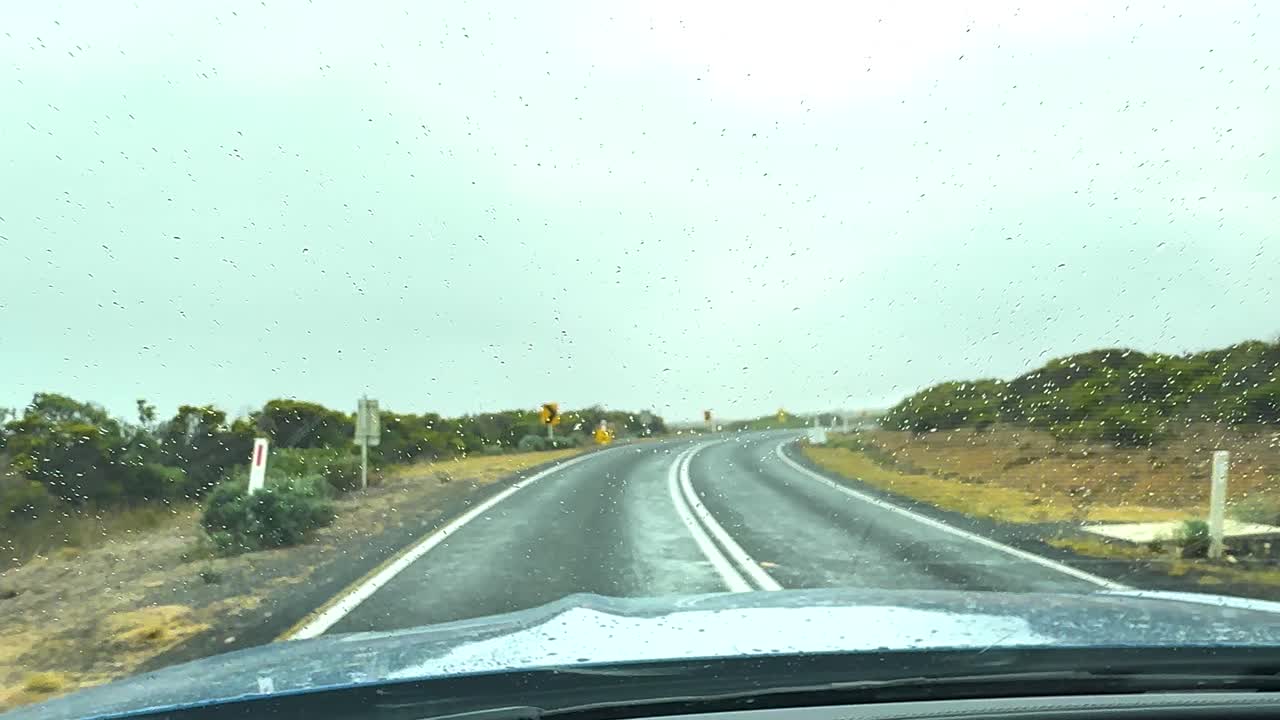 A car travels on a rainy day along the scenic Great Ocean Road in Victoria, Australia, with windshield wipers in motion