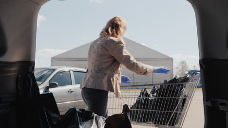 a woman in a mask and gloves puts shopping bags in the trunk of a car