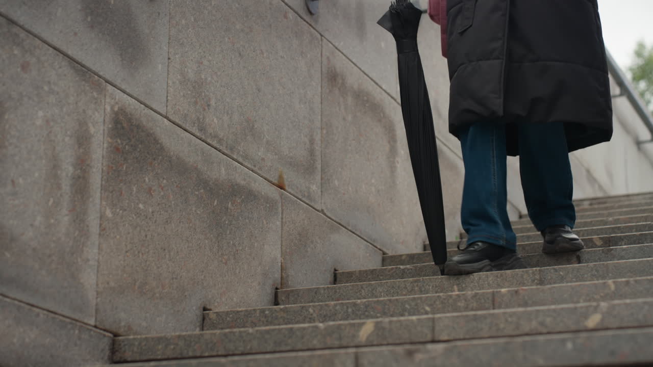 woman walking down concrete stairs holding closed umbrella, wearing long dark coat and jeans, descending stone stairway in overcast city, hand on metal handrail, closeup feet shots and umbrella tip