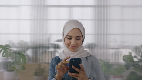 Porträt-Einer-Jungen-Muslimischen-Geschäftsfrau,-Die-Im-Hintergrund-Eines-Büroarbeitsplatzes-Mit-Einem-Smartphone-SMS-Schreibt-Und-Online-Surft