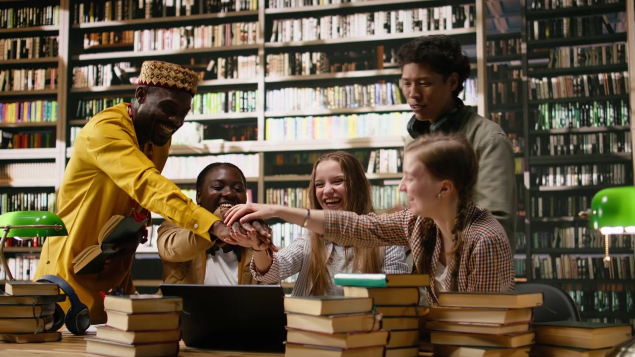 Smiling diverse students stacking hands together in unity at library table