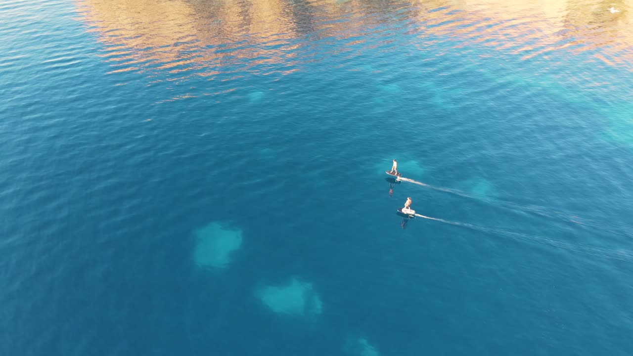 전기 서핑보드를 타고 칼라 에스콘디다 해안선에서 바다를 가로질러 서핑하는 두 사람 뒤의 공중 전망