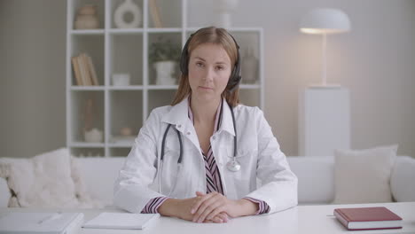 young female physician is listening patient or colleague by headphones using video calling nodding to camera