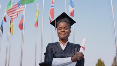 una joven graduada afroamericana sonriente de pie frente a la cámara con un diploma en las manos. la estudiante está afuera con las banderas internacionales en el fondo.