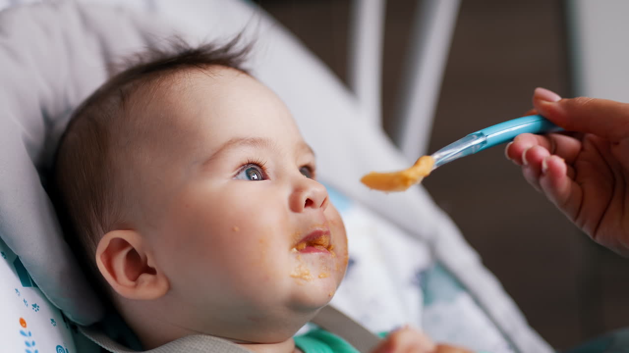 Healthy little baby boy being active during meal. Mommy gives her son some food and child rises hands and waves them.