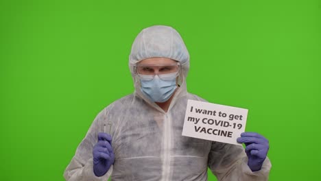 a man in a protective suit holding a syringe and a sign that says 'i want to get my covid-19 vaccine'