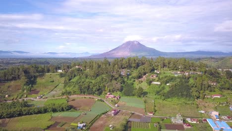 panning drone shot of mount sinabung surrounded by green village, pine trees, forest, and clouds in berastagi, north sumatera indonesia