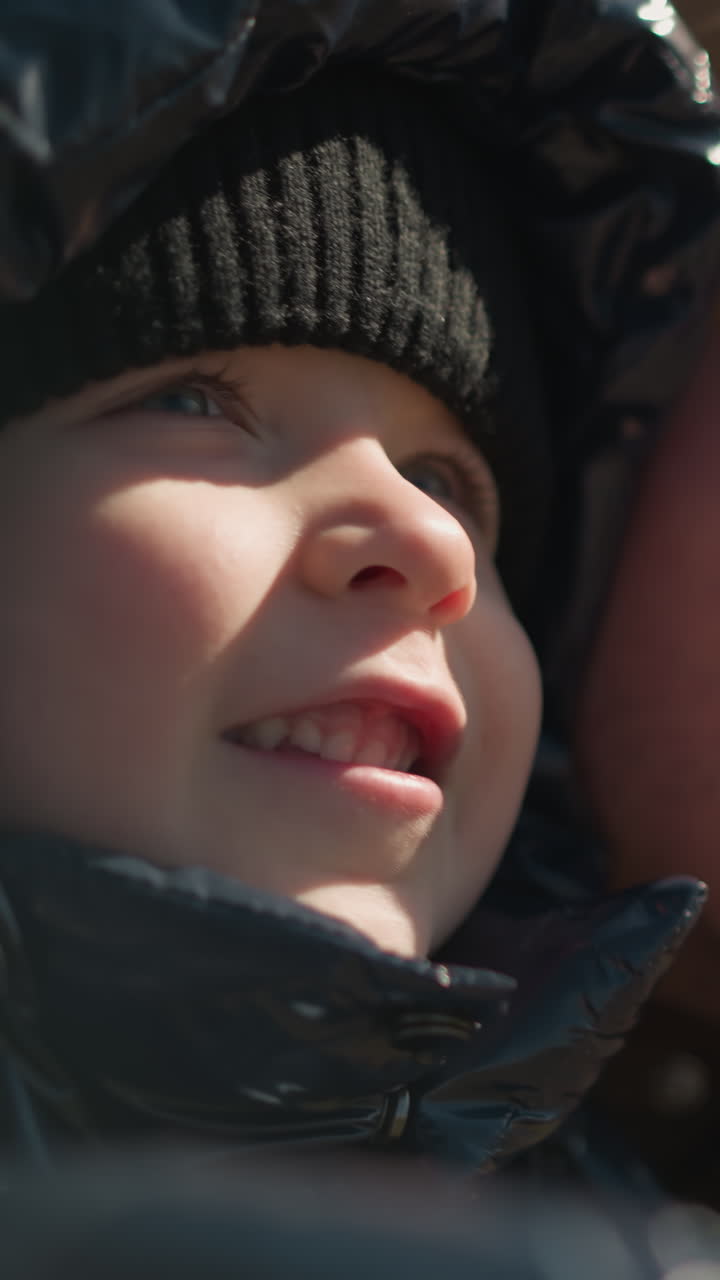 A close-up of a father and his son smiling and looking upwards with joy, the child is dressed in a shiny black jacket and beanie, while the father wears a brown jacket and a beanie
