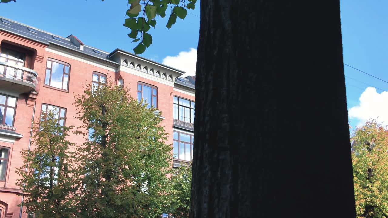 Charming red brick residential building with large windows and rooftop features, framed by lush green trees and a bright blue sky on a sunny day in the city