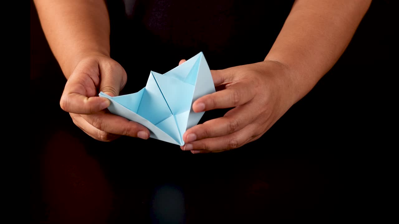 Person folds blue paper into origami boat under studio lighting, close-up, black background, steady camera