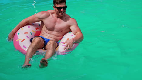 sportive caucasian man sitting in the swimming circle in the pool. man in sunglasses tries to row with hands.