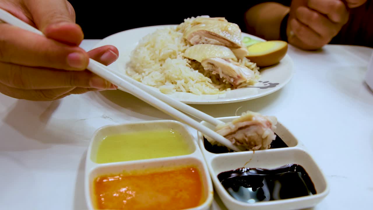 Close-up of hand using chopsticks to dip Hainanese chicken rice into sauces on white table