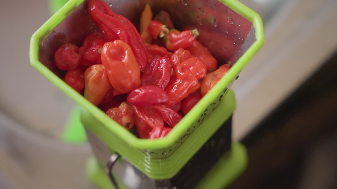 Primo piano di un tritapepe pieno di peperoni, una mano che inserisce i baccelli in un macinino manuale verde, peperoncini rossi vivaci che brillano di succhi, semi visibili, preparazione sensoriale di spezie sul piano di lavoro, cucina intima.