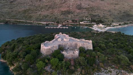 Aerial-orbit-view-of-empty-Ali-Pasha-Castle-with-golden-sunrise-over-huge-mountains-in-Porto-Palermo-bay,-Albania