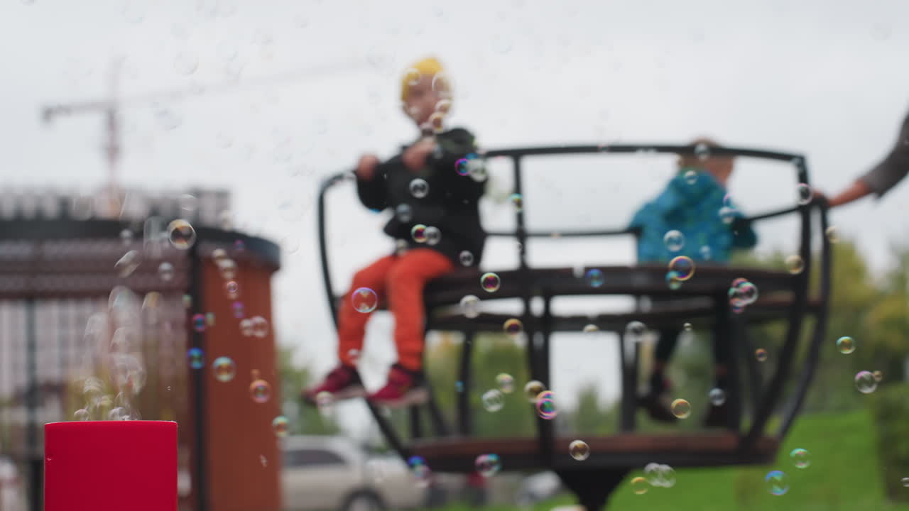 Foreground bubbles drift from red machine while mother spins two children on merry go round, soft focus park with gazebo and crane behind, cloudy sky, green lawn and playful motion