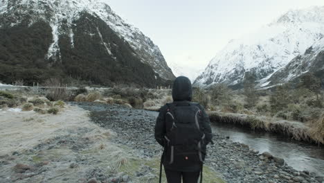 Mädchen-In-Wanderausrüstung-Spaziert-Am-Fluss-Entlang,-Umgeben-Von-Schneebedeckten-Bergen