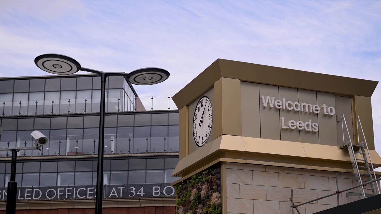 Welcome to Leeds sign and clock tower stand beside a modern glass office block under a soft daytime sky.