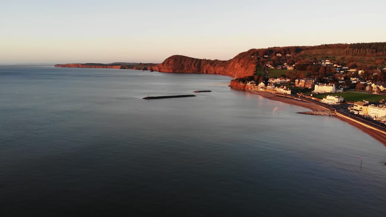 Aerial Along Sidmouth Coastline With Offshore Breakwaters Bathed In Golden Sunrise Light