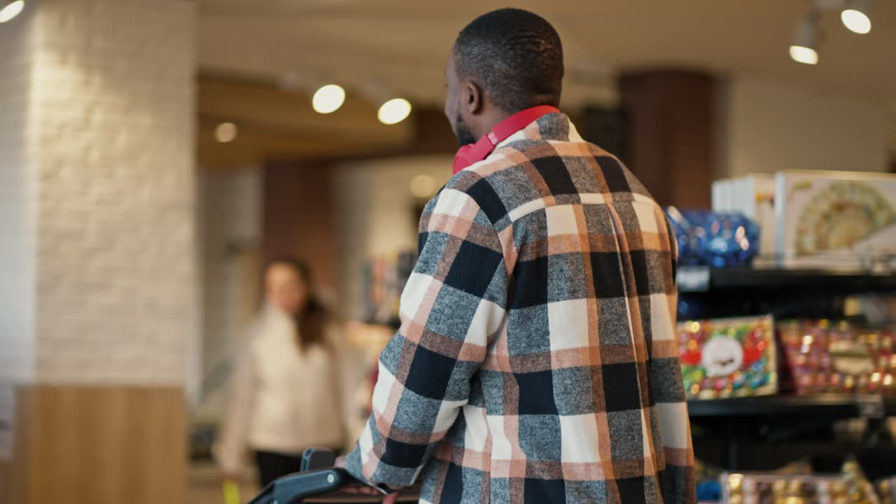 A man with Black skin in a plaid shirt and red wireless headphones walks through a modern grocery store and examines the goods on the counter