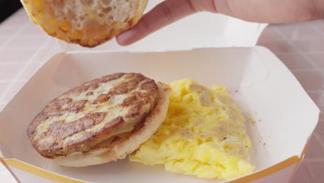 breakfast sandwich and omelet in cardboard tray, hand taking bread slice then placing it on vegan patty