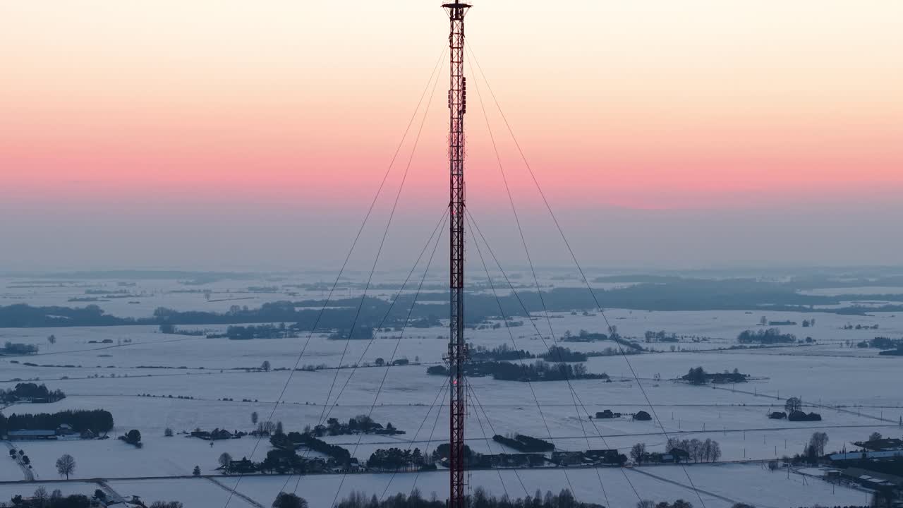 Video con drone che mostra un'alta torre di trasmissione in un vasto paesaggio innevato al tramonto. Le riprese aeree catturano la serena e ampia campagna agricola sotto un cielo caldo e colorato.