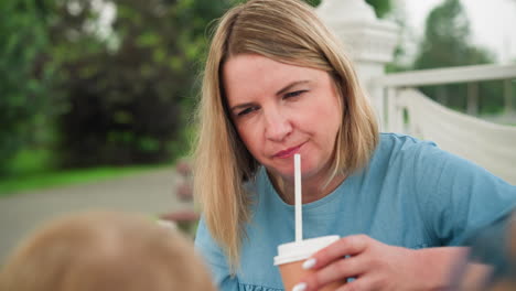 a woman drinks chocolate from a straw while sitting outdoors, looking focused, the back view of two children s heads is visible as they interact nearby