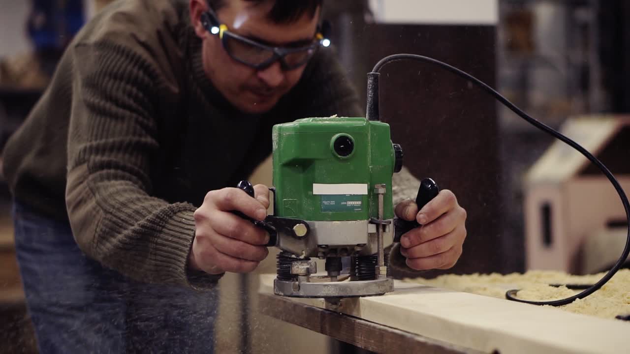 un maestro en gafas protectoras trabajando con una máquina de molienda manual en un taller de madera. moliendo una gran tabla de madera. polvo y astillas se esparcen por el suelo. cámara lenta