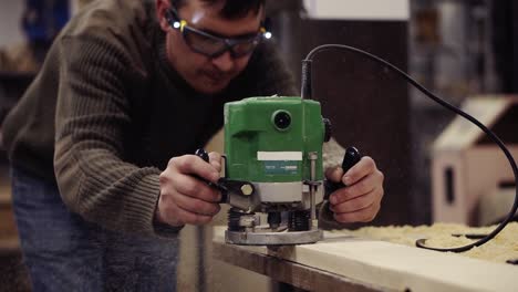 A-master-in-protective-glasses-working-with-manual-grinding-machine-at-wood-workshop.-Grinds-a-large-wooden-plank.-Dust-and-chips-are-scattering-on-the-floor.-Slow-motion