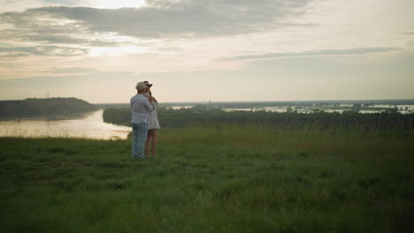 a romantic moment as a man in a hat, white shirt, and jeans embraces a woman in a white dress from behind. they stand together on a grassy hill beside a serene lake at sunset