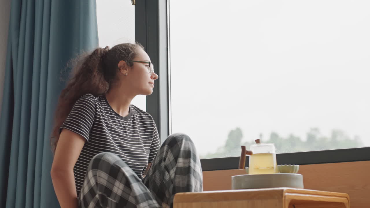 Donna alla finestra dell'hotel che si allunga per prendere la teiera, ospite nomade digitale che si prepara per la giornata mentre si ferma ad ammirare la vista sulla città, pantaloni lounge a quadri, tavolino in legno con teiera e tazza, interni tranquilli, luce soffusa.