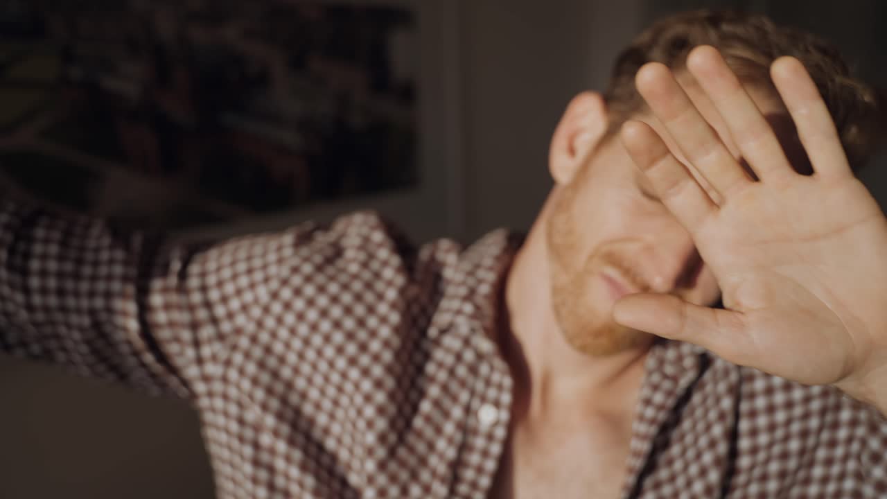 Man opens window blinds and shields eyes from bright sunlight, squinting and raising hand to block glare indoors. Morning light floods the room, creating strong shadows on his face. guy blinded by sun