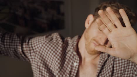 man opens window blinds and shields eyes from bright sunlight, squinting and raising hand to block glare indoors. morning light floods the room, creating strong shadows on his face. guy blinded by sun