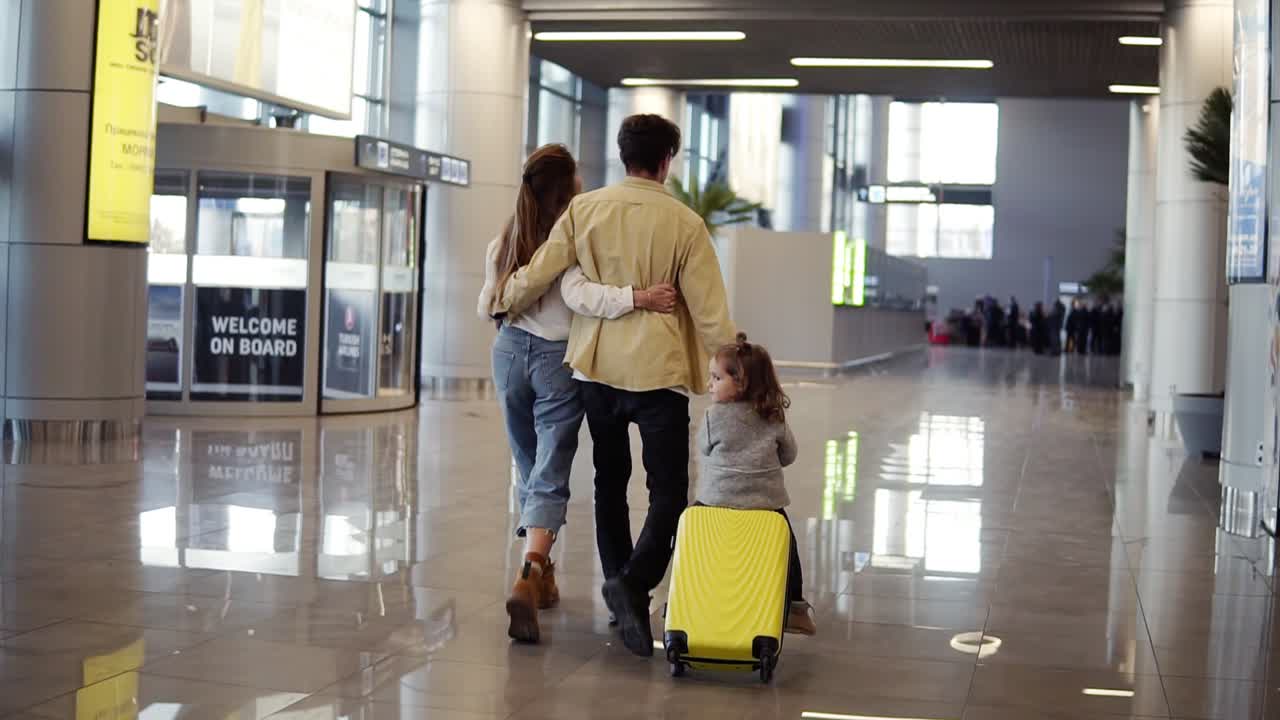 vista retrospectiva de la familia caucásica moderna - madre, padre, hija - caminando por el pasillo del aeropuerto y montando a su hija en una maleta antes de la salida a las vacaciones. cámara lenta