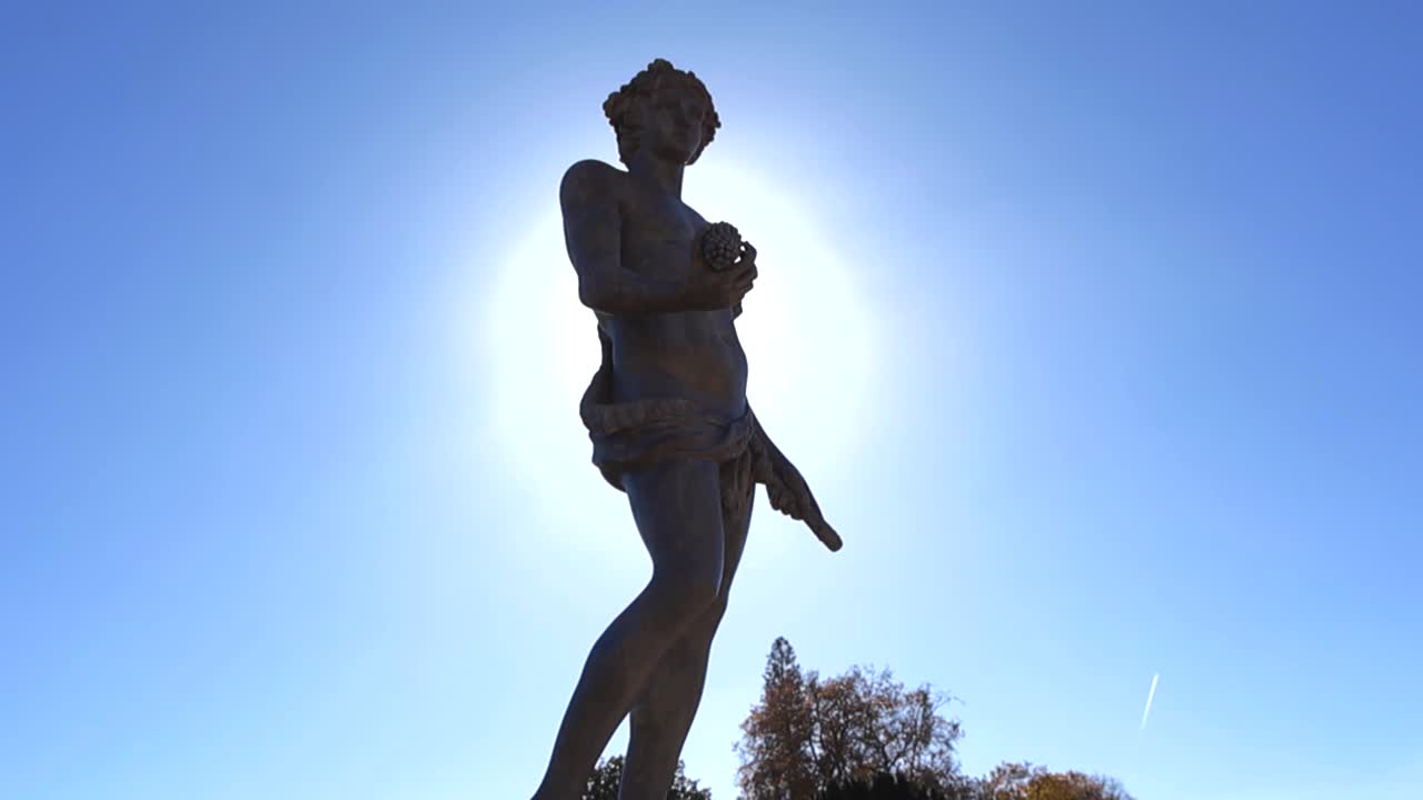 Statua femminile in pietra retroilluminata nei giardini di un palazzo, illuminata dalla luce del sole mentre la telecamera orbita, creando una silhouette drammatica contro un cielo limpido