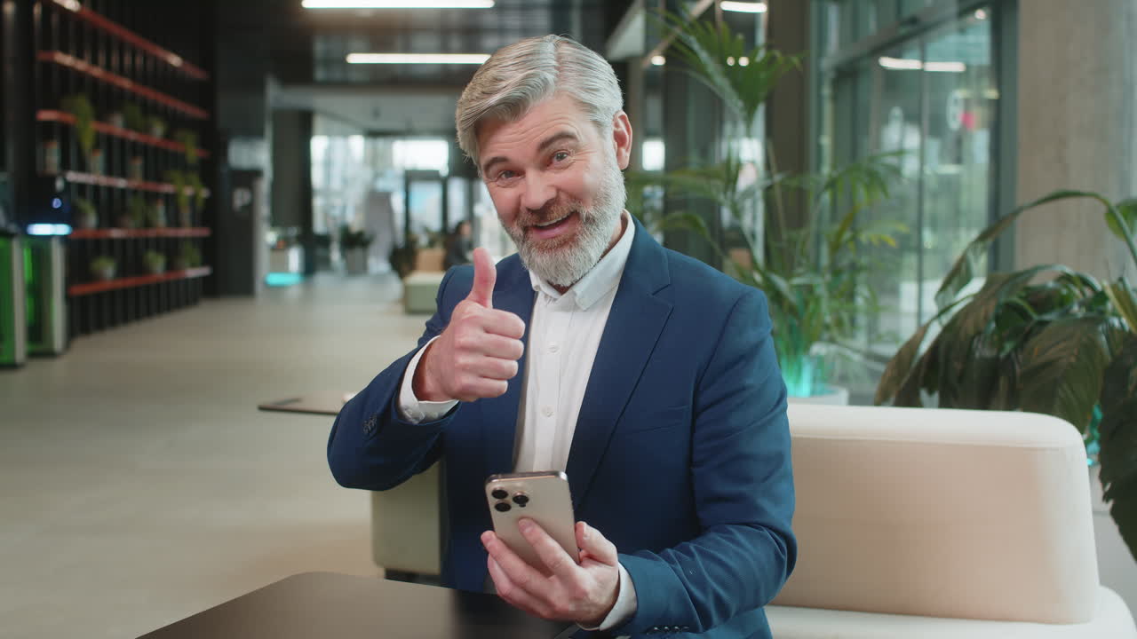 Smiling mature businessman in suit recommends using the phone app sitting in modern office lobby