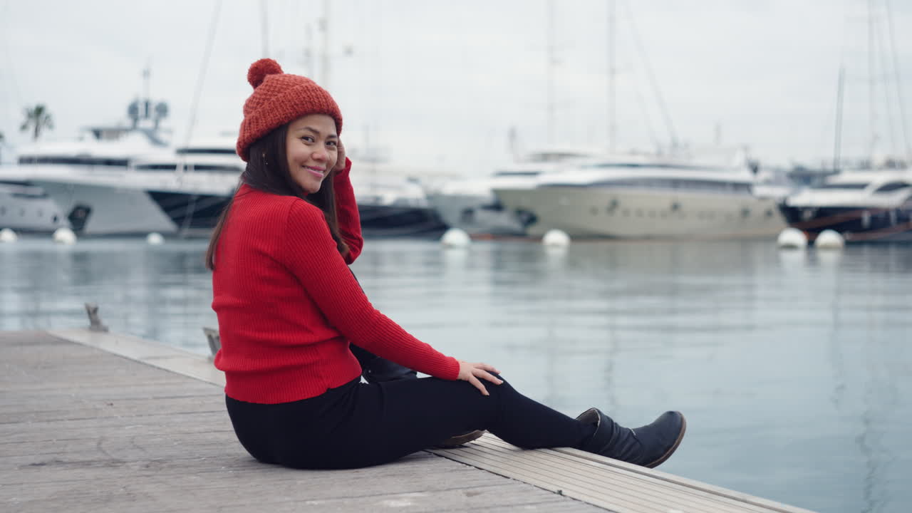 una mujer asiática sonríe a la cámara mientras se pone un gorro rojo, que coincide perfectamente con su camisa de cuello rojo, en un puerto con algunos yates como telón de fondo en el puerto de valencia, españa