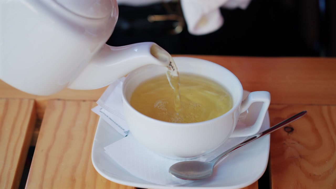 Tè caldo versato da una teiera bianca in una tazza di ceramica su un tavolo di legno.