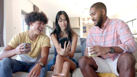 at home, diverse friends hanging out, holding mugs and smartphone, sitting on couch chatting