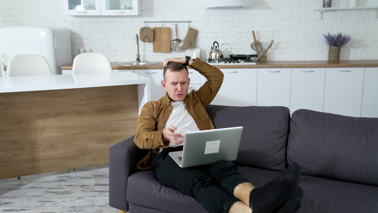Young man playing computer games at home. Nervous guy laying on a couch with a laptop on kitchen background.